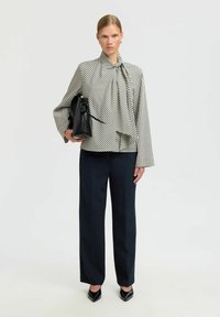 Kvinde iført en bluse med sort-hvide striber og en sløjfe, sorte videbukser, sorte spidse hæle og holder en sort lædertaske.