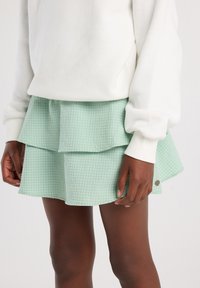 Mini falda verde de capas con un patrón texturizado de cuadros, que presenta un dobladillo deshilachado y un pequeño acento de logo metálico.