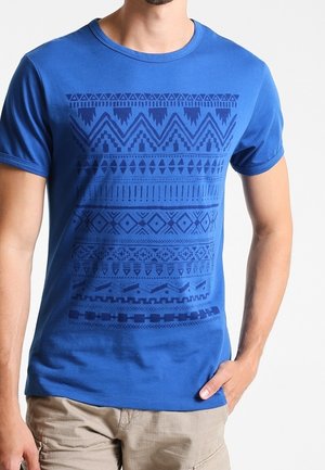 Mann trägt ein blaues kurzärmeliges T-Shirt mit dunkelblauen geometrischen Mustern und beigen Shorts, Hand ruht in der Tasche.