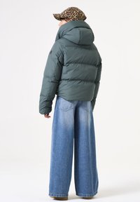 Chaqueta acolchada verde con un cuello alto; combinada con unos vaqueros de mezclilla azul de pierna ancha. La gorra tiene un patrón de leopardo.