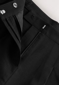 Pantalon noir à texture lisse, doté d'une fermeture éclair, d'un bouton en métal et d'un design à devant plat avec poches latérales.