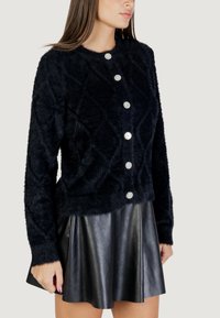 Veste en fausse fourrure noire avec un motif matelassé en losanges, dotée de cinq fermetures à boutons brillants et de longues manches, portée sur une jupe en cuir noire.