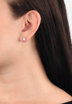 Pendientes de acero plateado con una gema redonda y clara rodeada de un halo texturizado. Llevados en la oreja, mostrando un primer plano.
