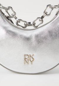 Srebrna metaliczna torebka o teksturowej powierzchni, z logo "DKNY" w kolorze złotym i łańcuszkiem z przeplatającymi się ogniwami.