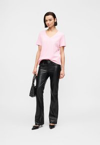 Γυναίκα που φοράει ροζ T-shirt με V λαιμόκοψη, μαύρο δερμάτινο παντελόνι, μαύρα μυτερά παπούτσια και κρατάει μια μαύρη τσάντα με καρφιά.