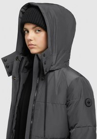 Gray insulated jacket with a hood, featuring snap and zipper closures, quilted texture, and a round logo patch on the sleeve.