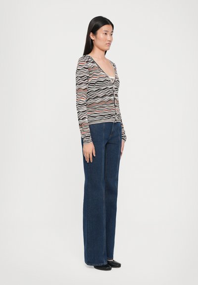 Cardigan a righe ondulate nei toni del nero, rosa e crema, con scollatura a V profonda e chiusura con bottoni; abbinato a jeans a zampa d'elefante blu.