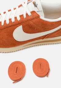 Baskets Nike en daim orange avec swoosh blanc et lacets, accompagnées de deux lacets orange roulés sur fond blanc.