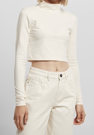 Kvinde iført langærmet cremefarvet crop top og cremefarvede bukser med høj talje, som viser en navlepiercing mod en ensartet baggrund.