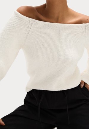 Femme portant un pull en maille blanc à épaules dénudées et un pantalon noir, main posée sur la hanche, sur un fond clair uni.