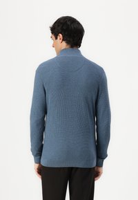 Blue textured sweater with a high collar, long sleeves, and a straight hem. Worn with dark trousers, the back design features a smooth finish.