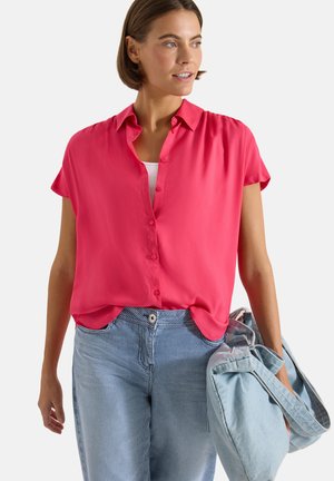 Femme portant une chemise boutonnée à manches courtes rose vif et un pantalon bleu à fines rayures, tenant un grand sac en tissu bleu clair à ses côtés.