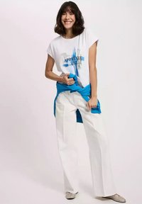 Biała bawełniana koszulka z niebieskim graficznym napisem, połączona z luźnymi białymi spodniami oraz jasnoniebieskim swetrem przewiązanym w talii. Teksturowane buty.