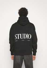 Sort hættetrøje med snor, med stor hvid tekst "STUDIO" på ryggen. Fremstillet af et blødt stof med ribkant ved ærmer og bund.