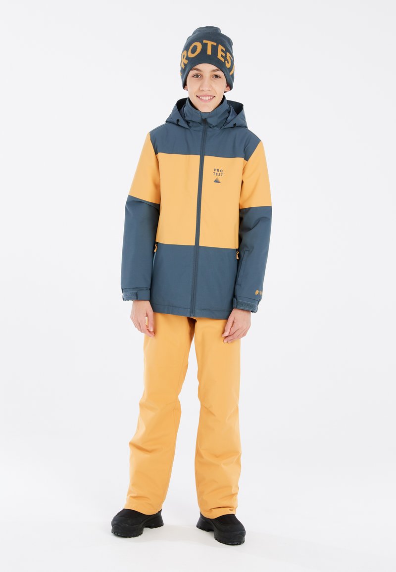 Jungen Winterjacke in Blau und Orange mit Kapuze, ausgestattet mit einem Frontreißverschluss und Logo. Kombiniert mit orangefarbenen wasserfesten Hosen und schwarzem Schuhwerk.