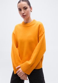 Πορτοκαλί μάλλινο πουλόβερ με ριμπ λαιμόκοψη και μανσέτες, με άνετη εφαρμογή και μακριά μανίκια. Συνδυάζεται με μαύρο παντελόνι.