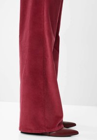 Pantaloni in corda rossa, larghi e con righe verticali, abbinati a delle scarpe con punta bordeaux. Il tessuto mostra una sottile trama e una profondità di colore.