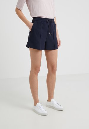 Kvinde iført marineblå shorts med snøre, lyserød kortærmet top og hvide sneakers, stående med den ene hånd i lommen mod en ensfarvet baggrund.
