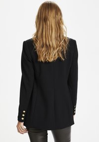 Černý blazer se strukturovanými rameny a třemi zlatými knoflíky na každém rukávu. Materiál vypadá hladce a má dobře padnoucí střih.