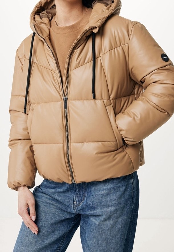 Personne portant une doudoune matelassée beige avec des cordons noirs par-dessus un pull beige et un jean bleu, les mains dans les poches.