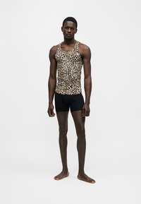 Homme debout pieds nus, portant un débardeur imprimé léopard et un short noir ajusté, sur un fond uni.