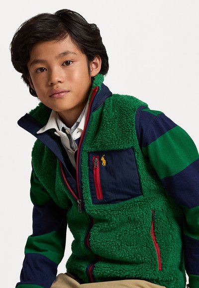 Γυμναστική γιλέκο με πράσινη, υφασμένη βάση, ναυτικές μπλε μανίκες με ρίγες, κόκκινες λεπτομέρειες στον φερμουάρ και ένα μικρό κίτρινο λογότυπο στην τσέπη του στήθους.