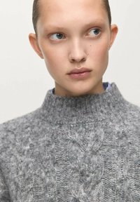 Szary sweter w sploty z wysokim kołnierzem. Posiada fakturowany wzór, prążkowany dekolt i miękki, wełnopodobny materiał.