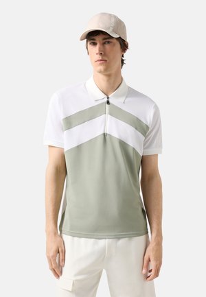 Άνδρας που φοράει λευκό και ανοιχτό γκρι πόλο με κοντό μανίκι και φερμουάρ, με μπεζ καπέλο, στέκεται μπροστά σε απλό ανοιχτό φόντο.