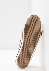 Semelle d'une basket rouge avec un motif ondulé et le nom de la marque embossé, placée verticalement contre un angle de mur blanc.