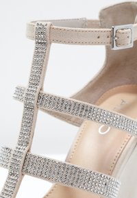 Sandale à talons hauts en daim beige avec des sangles argentées cloutées, une boucle de cheville et une doublure intérieure lisse. Accentuée par un talon en wedge.