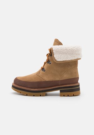 Bottine en daim beige avec semelle en caoutchouc marron, fermeture à lacets sur le devant, col en polaire blanche rabattable et petit talon carré sur fond blanc.