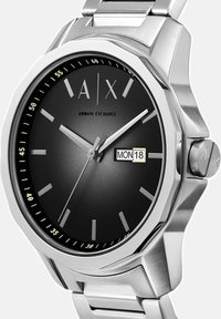 Kovinsko jekleno uro s črnim številčnikom, oktagonalno ohišje, srebrni kazalci in prikaz datuma. Ima kovinsko zapestnico s sponko.