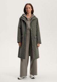 Cappotto lungo trapuntato verde oliva con cappuccio, dotato di tasche laterali e chiusure a scatto, indossato sopra un completo grigio e scarpe da ginnastica bianche.