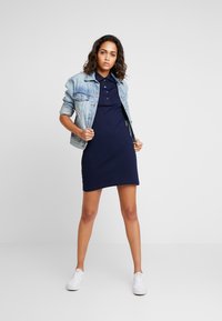 Navy polo mekko, jossa on lyhyet hihat, slim fit -malli ja sivuroikkuja, kuitenkin yhdistettynä vaaleansiniseen farkkutakkiin ja valkoisiin tennareihin.