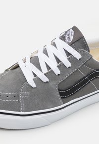 Sapatilha de camurça cinza com atacadores brancos, detalhes pretos e um patch do logótipo na língua. Apresenta uma superfície texturizada e sola de borracha branca.