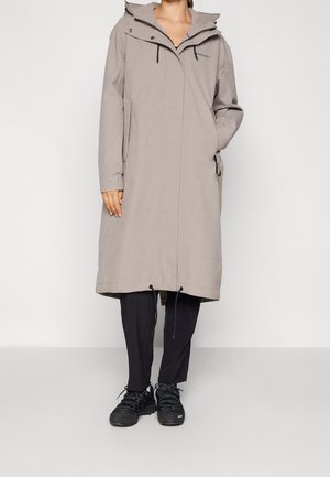 Personne portant un long manteau beige à capuche sur un pantalon noir et des baskets noires, debout devant un fond clair uni.