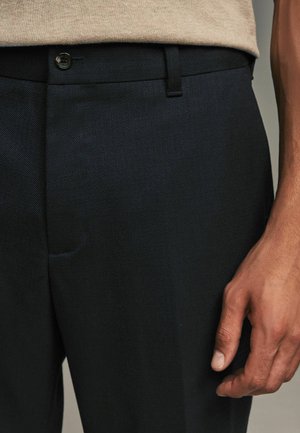 Pantalons bleu marine foncé en tissu texturé, dotés d'une fermeture à bouton, de passants de ceinture et d'une poche latérale. Gros plan sur la taille et la main.