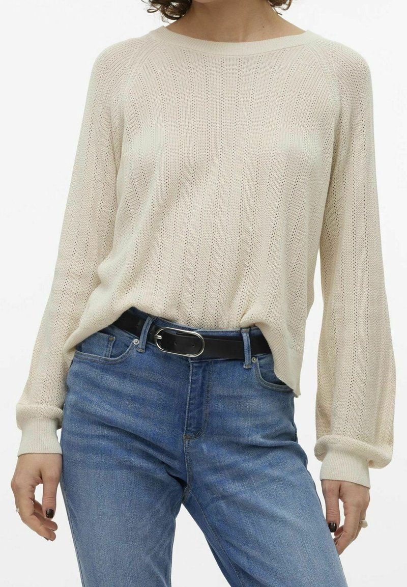 Femme portant un pull en tricot crème à manches longues rentré dans un jean bleu taille haute avec une ceinture noire, les mains détendues sur les côtés.