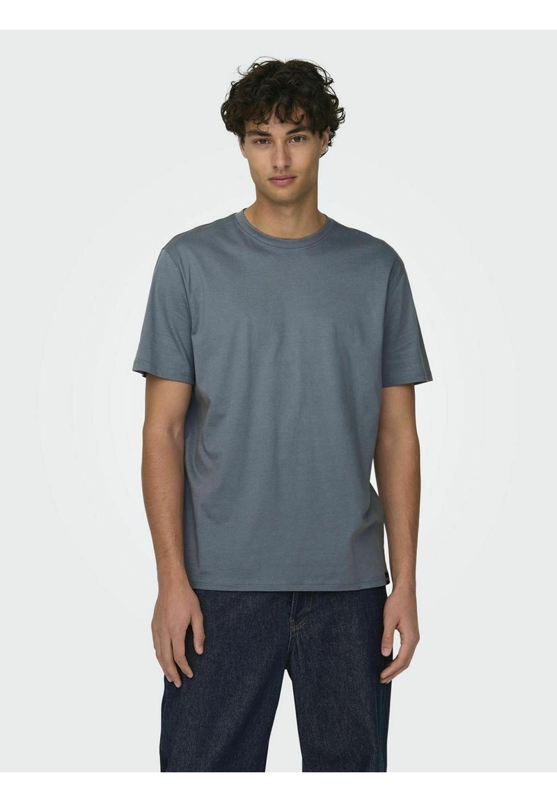 T-shirt grigia a maniche corte, realizzata in cotone, con scollatura rotonda e vestibilità regolare, abbinata a jeans scuri. Design semplice, senza motivi visibili.