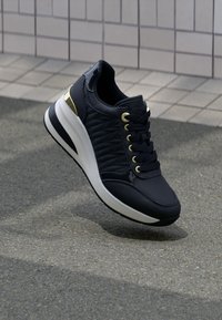 Schwarze Sneaker mit goldenen Ösen und Fersendetails, weißer und schwarzer Sohle, die über einem grauen Bürgersteig und einem gefliesten Wandhintergrund schwebt.