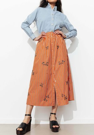 Femme portant un chemisier bleu à rayures boutonné, rentré dans une jupe midi orange à rayures verticales avec broderie florale et fente avant, associé à des sandales plateformes noires.