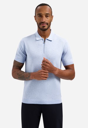 Man met een getatoeëerde arm die een lichtblauw poloshirt met korte mouwen en een ritskraag draagt, met zijn handen voor zich samengehouden.