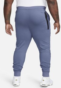 Blauwe sportieve joggings met een aansluitend ontwerp, enkelboorden en een zijzak met rits. Gemaakt van zacht, rekbaar materiaal. Draagt met witte sneakers.