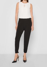 Biała bezrękawnik z dekoltem w kształcie łezki i ozdobami przy szyi, w połączeniu z czarnymi zwężającymi się spodniami i czarnymi szpilkami.