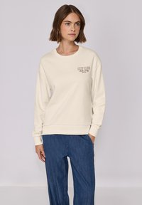 Felpa crewneck beige chiaro realizzata in tessuto morbido, con un piccolo logo "CITY CLUB" marrone sul petto e polsini a coste.