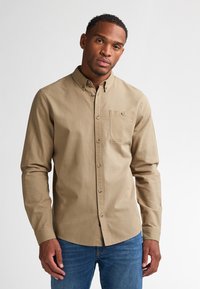 Langarmige beige button-up shirt met een borstzak en bruine knopen, gecombineerd met blauwe denim jeans, gepresenteerd op een model.