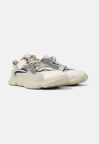 Beige mesh sneakers with grey and black accents, featuring thick rubber soles, textured overlays, and curved design lines for support.