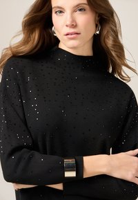 Czarny sweter dzianinowy z wysokim dekoltem, ozdobiony rozrzuconymi kwadratowymi aplikacjami i długimi rękawami. Srebrny akcent przy mankietach.