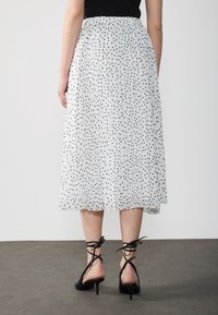 Falda blanca con pliegues y lunares negros, con cinturilla elástica, que cae hasta la mitad de la pantorrilla, combinada con zapatos de tacón negro con ataduras en el tobillo.