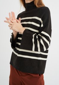 Suéter negro de punto con rayas horizontales blancas, cuello alto y mangas acampanadas, combinado con pantalones color óxido. Textura suave, diseño informal.